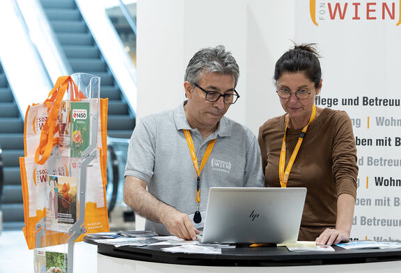 Eine FSW-Beraterin und ein FSW-Berater stehen am Beratungsstand im Einkaufszentrum Donauzentrum und blicken beide in einen Laptop