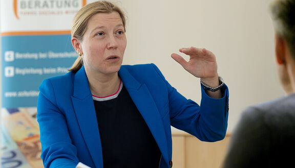 Gudrun Steinmann von der FSW Schuldenberatung sitzt an einem Tisch und ist in einem Gespräch mit einer männlichen Person während ihr linker Arm leicht angehoben ist