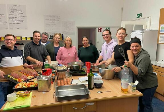 Eine Gruppe von 9 freiwilligen Frauen und Männen steht in der Küche der FSW Obdach-Wärmestube Sautergasse mit Küchenutensilien und Lebensmitteln.