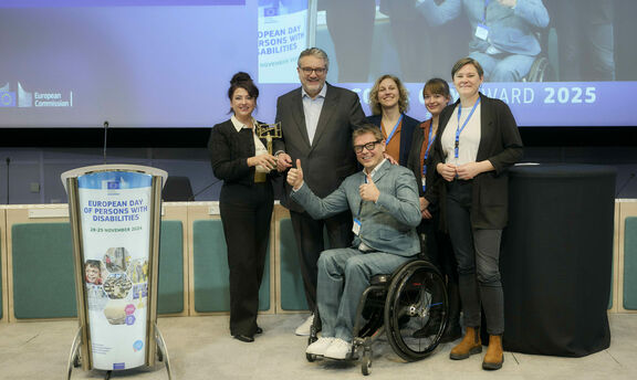 Eine Gruppe von Personen überreicht Stadtrat Peter Hacker auf einer Bühne den Access City Award 2025. Alle Anwesenden lächeln in die Kamera.