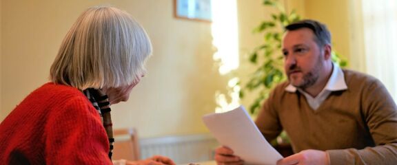 FSW Community Nurses Beratungsgespräch | FSW Ein Mitarbeiter berät eine ältere Dame wobei beide zu Tisch sitzen und er Dokumente in seiner Hand hält.