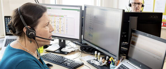 Mitarbeiterin von 1450 | FSW Eine Mitarbeiterin und ein Mitarbeiter von 1450 mit Headsets sitzen einander gegenüber und arbeiten an Computern in einem Büro.
