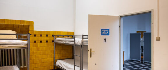 Nachtquartier Obdach Sautergasse | FSW Ein schlichtes Zimmer im Nachtquartier Obdach Sautergasse mit Stockbetten und einer teilweise gefliesten Wand. Eine offene Tür führt zu Duschen und Toiletten.