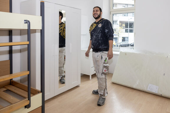 Ein jüngerer Mitarbeiter des Facility Management steht mit einem offenen Lächeln in einem Zimmer neben einem Kleiderschrank mit Spiegel und blickt in die Kamera. Links vom Bild sieht man ein leeres Stockbett und hinter dem Mitarbeiter sieht man ein Fenster und daneben eine an eine Wand angelehnte Matratze.