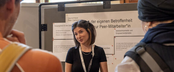 Infostation exPEERience | FSW Eine junge sprechende Frau steht vor einer Pinnwand mit einem Plakat mit der Aufschrift: von der eignen Betroffenheit zur Peer-MitarbeiterIn. Einige Personen hören ihr zu.
