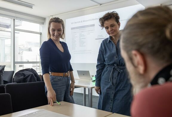 Katharina Kotynkowiecz steht neben Simone Einfalt-Schneider vor einem Tisch. Beide schauen lächelnd zu den am Tisch liegenden Unterlagen.